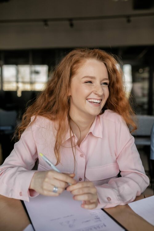 Smiling woman with red hair in a pink blouse, taking notes, embodying the creative and user-friendly experience of building a WordPress website, as endorsed by top industry players.